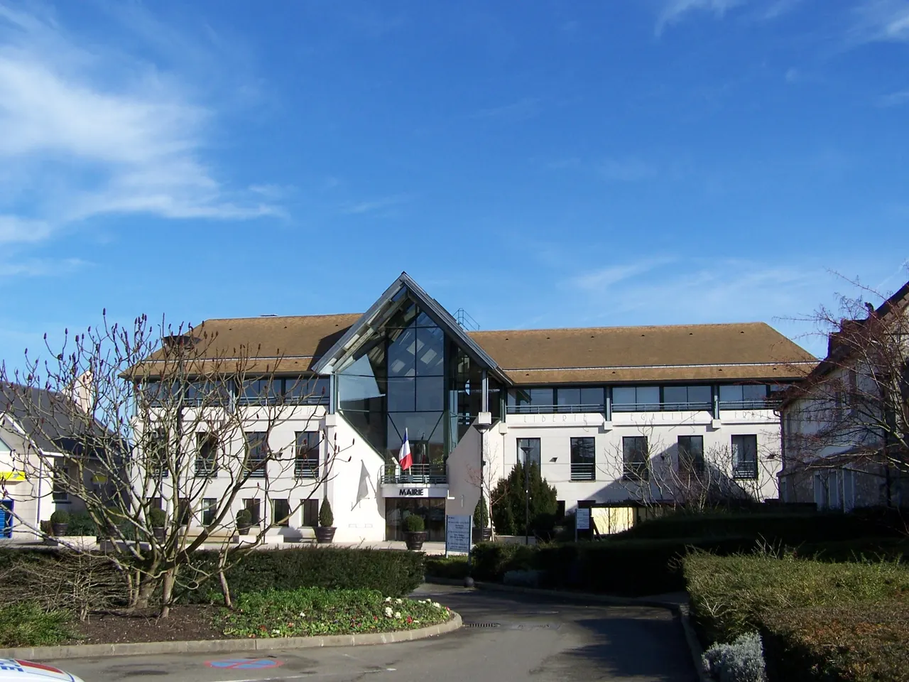 L’hôtel de ville de Voisins-le-Bretonneux, sa façade blanche et son entrée vitrée en pignon, derrière des massifs et un magnolia sans feuilles.